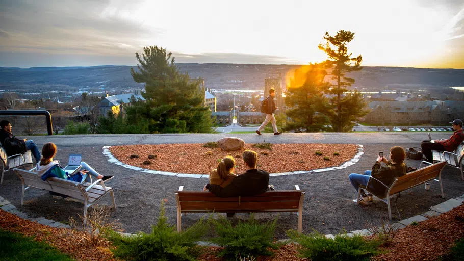 Two students are sitting on a bench, seen from behind, watching the sun set on the distant horizon.