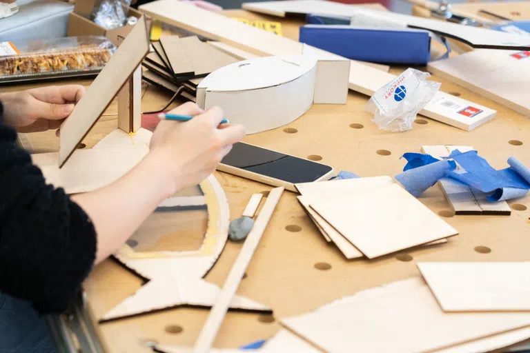 Closeup of a table full of various cardboard cutouts and other art-project materials. At left you can see a hand holding a blue pencil. 