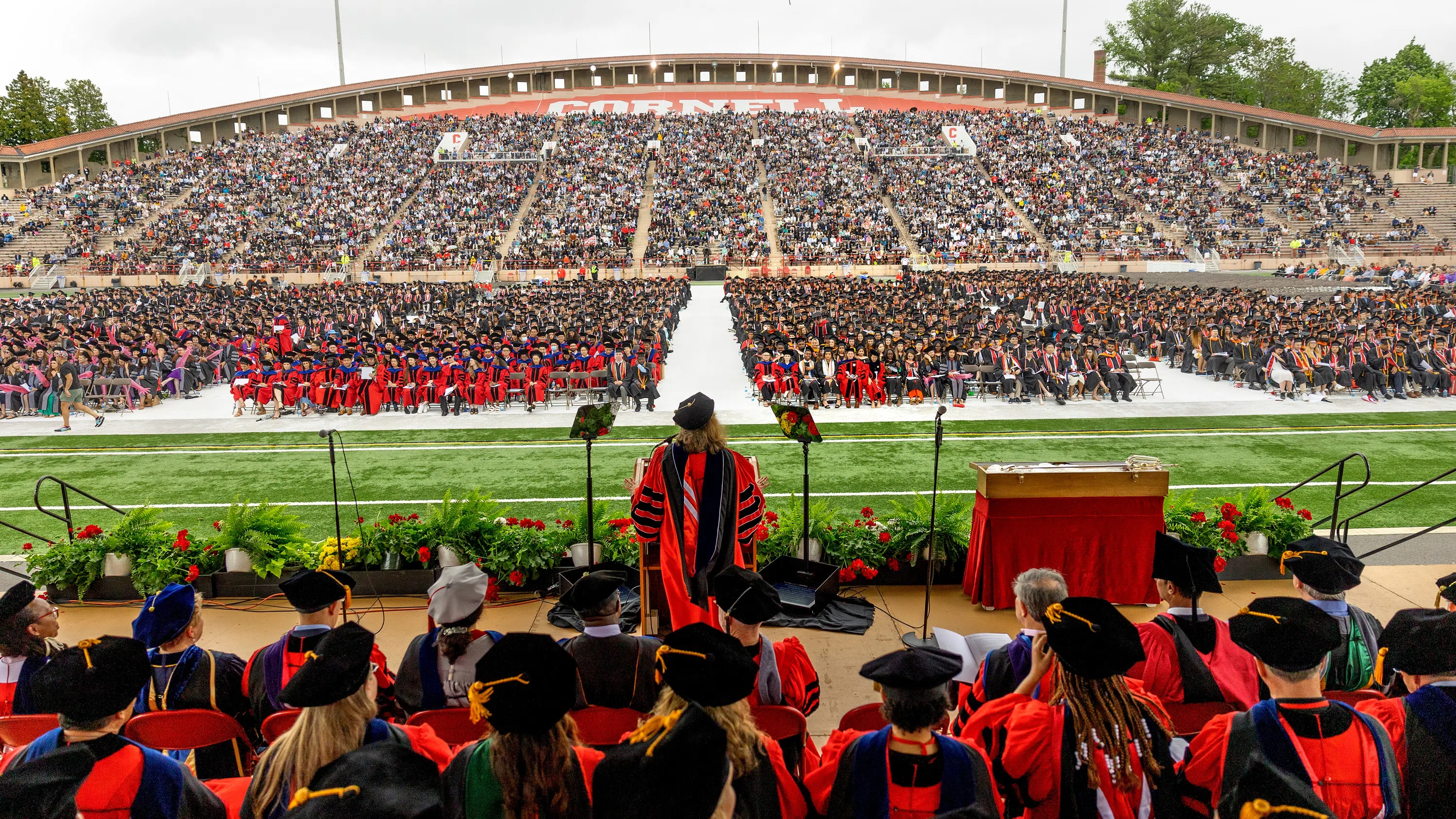 Cornell University president Martha E. Pollack, seen from behind dressed in academic regalia, addresses a crowd of thousands at Schoellkopf Field. Behind her are several more individuals, also wearing regalia.