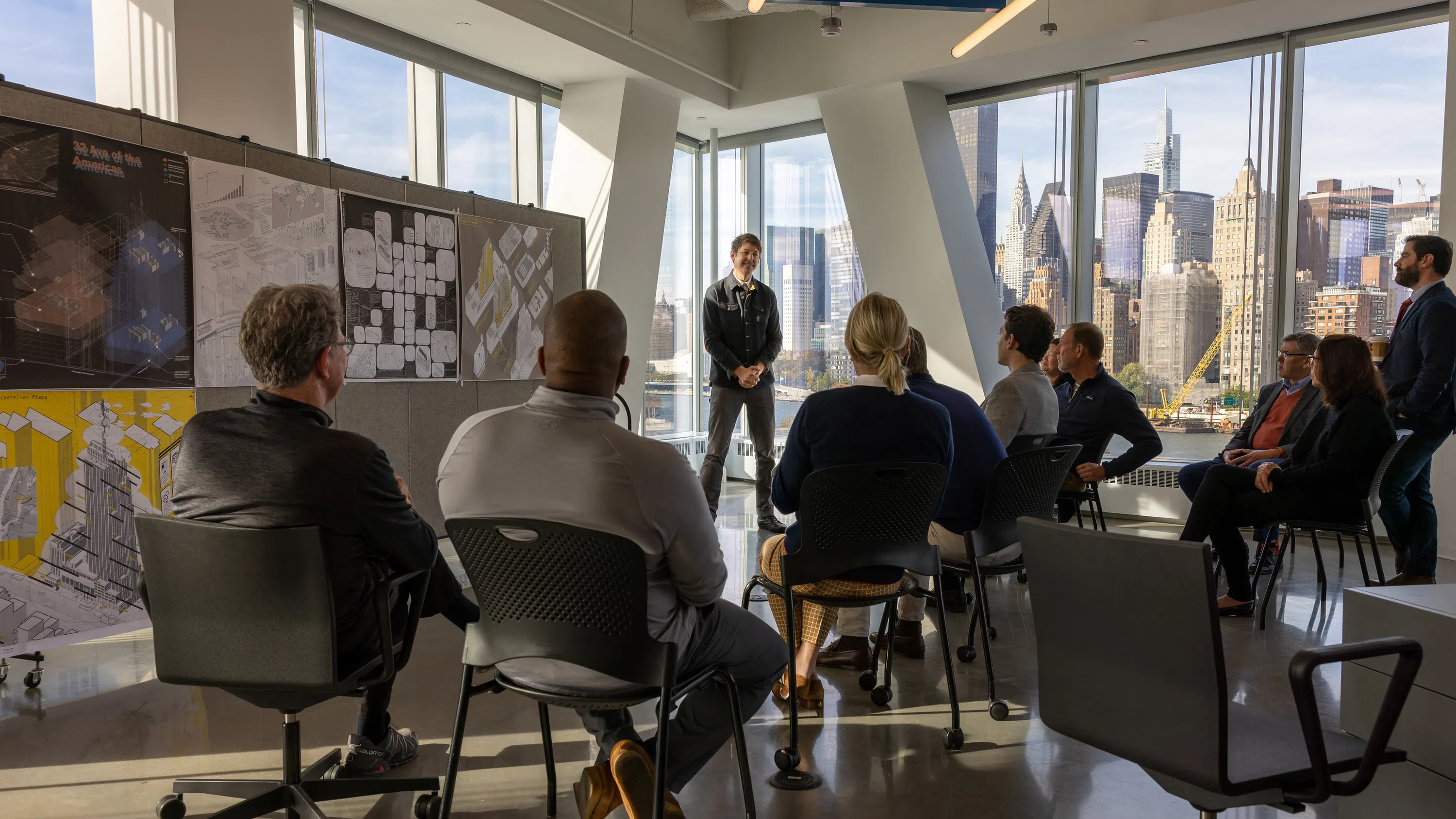 About a dozen people sitting in chairs, seen from behind, watching someone standing and speaking to them. Behind them is the skyline of Manhattan, including the Chrysler Building.