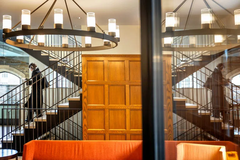 A student descends the stairs in Myron Taylor Hall with a chandelier hanging above, while the same image is reflected in a nearby window.