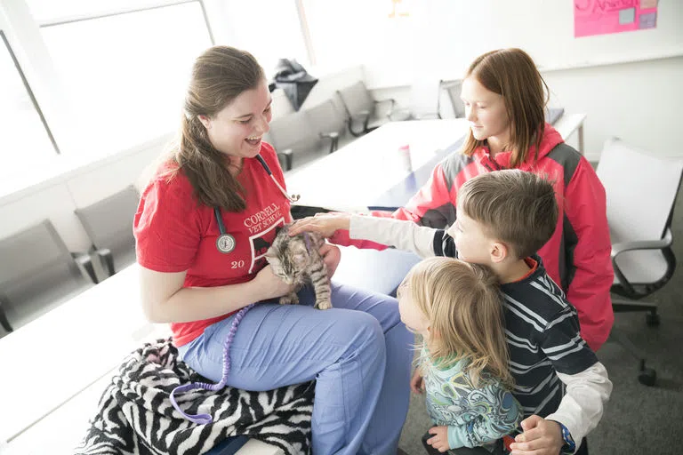 A Cornell Vet student sits and holds a kitten while three children eagerly reach out to pet it during the annual College of Veterinary Medicine Open House event.