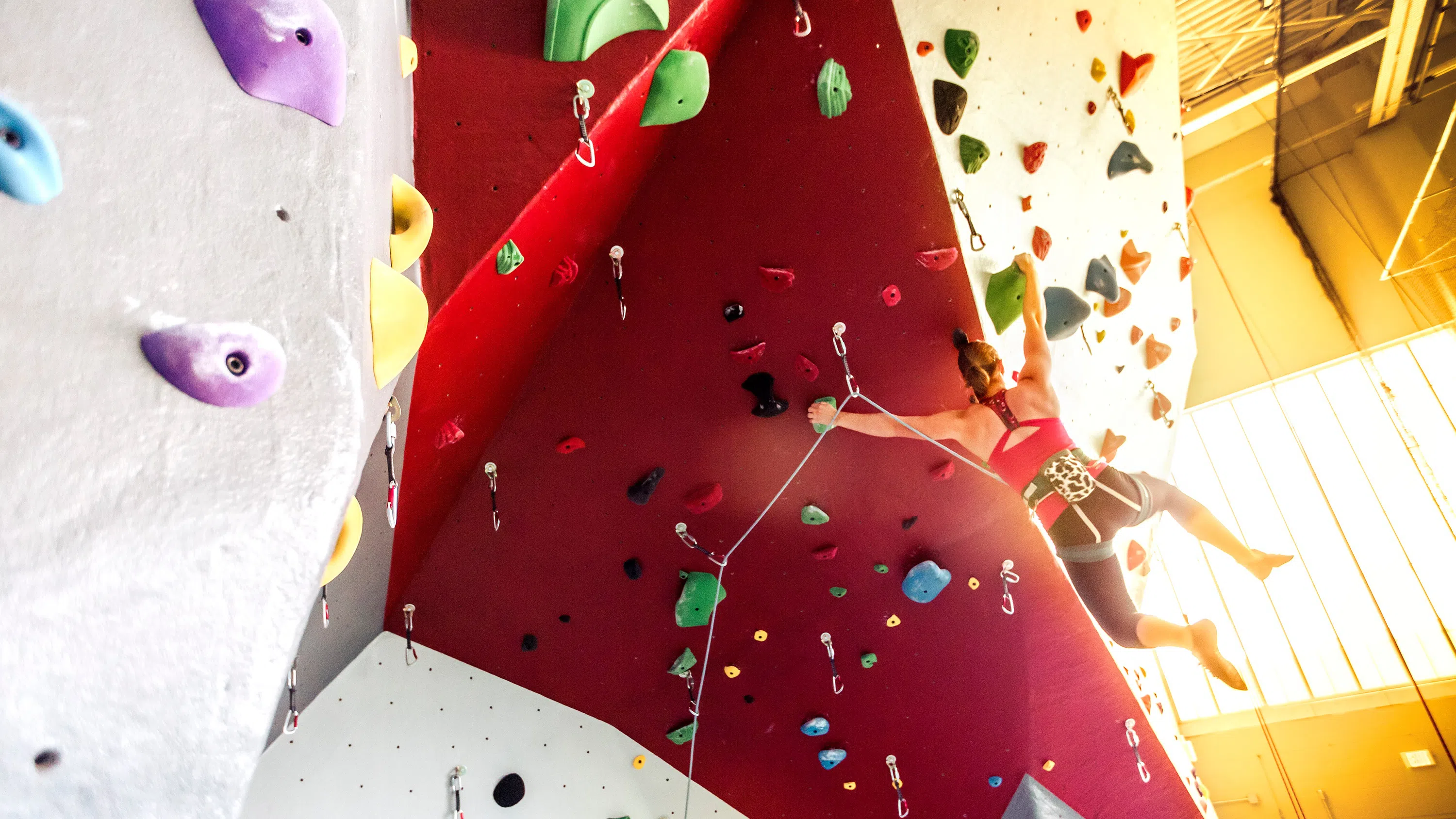 A student scales the Lindseth climbing wall. They’re hanging onto a wide, red-colored portion of the climbing wall, which is studded with dozens of handholds.