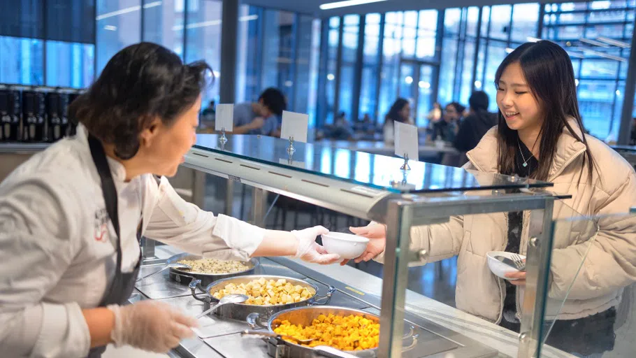 A Cornell Dining staff member hands a bowl to a customer. Between them are three large containers of buffet-style food.