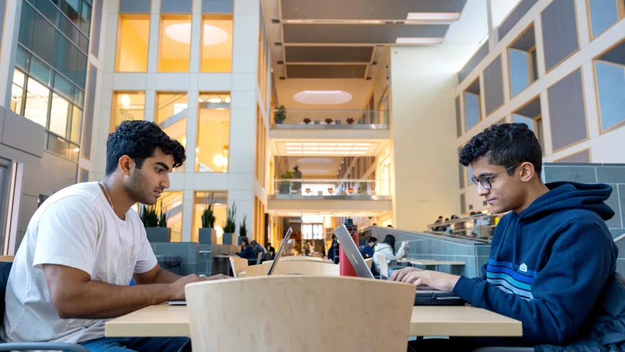 Two students, studying, facing each other at a table in a large, airy atrium.