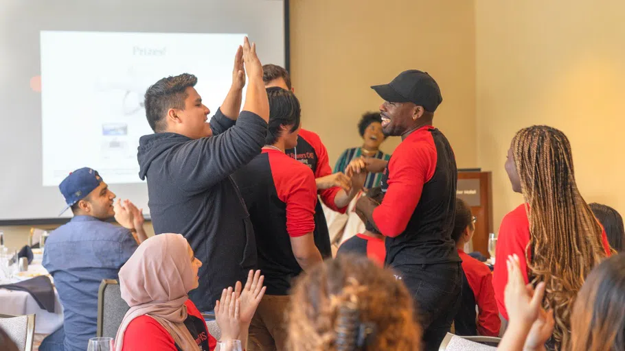 Two students, standing at center, smile and give each other high-fives while others, seated, clap and look on.