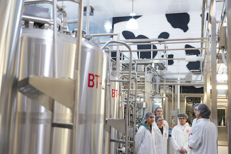 Students wearing white lab coats and plastic hair coverings stand among large stainless steel machines, learning how ice cream is manufactured in the Cornell dairy facility in Stocking Hall.