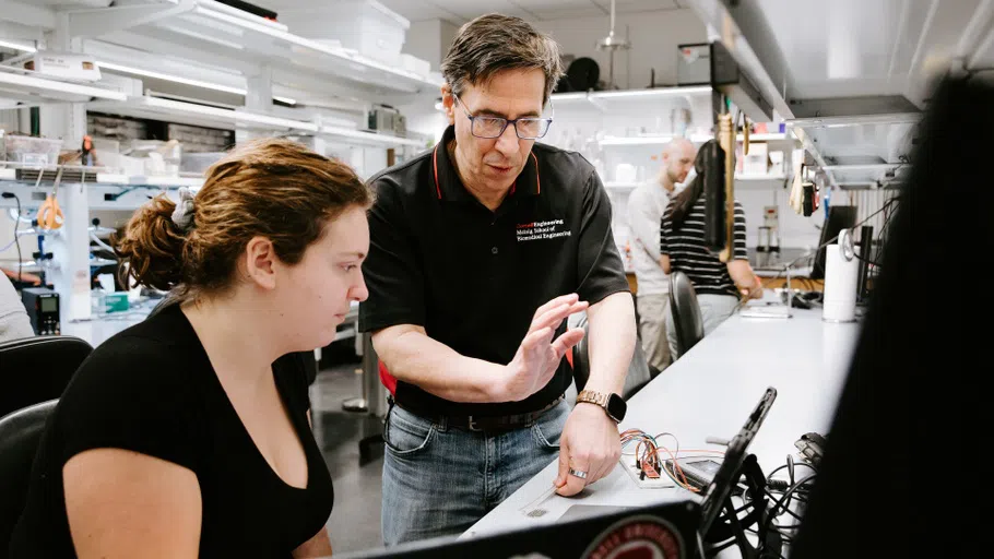 A faculty member, standing, guides a student on a biomedical engineering project.