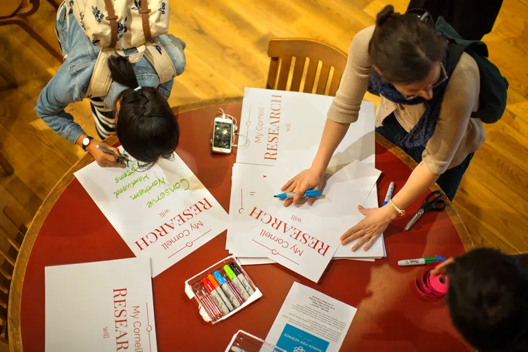 A closeup overhead shot of a table, with students using colorful markers to write on posters that say, “My Cornell Research will…” at the Big Red Barn.