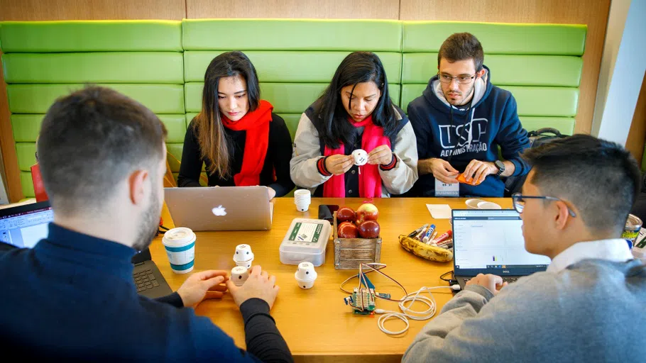 Five students sit at a table, upon which are several laptops, small pieces of electronic equipment, and other items as they work on a project.