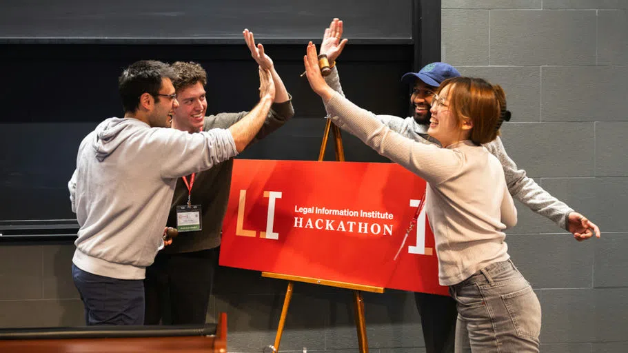 Four students of the winning team celebrate their victory with high fives over a red “Legal Information Institute Hackathon” sign during the inaugural event in Myron Taylor Hall.