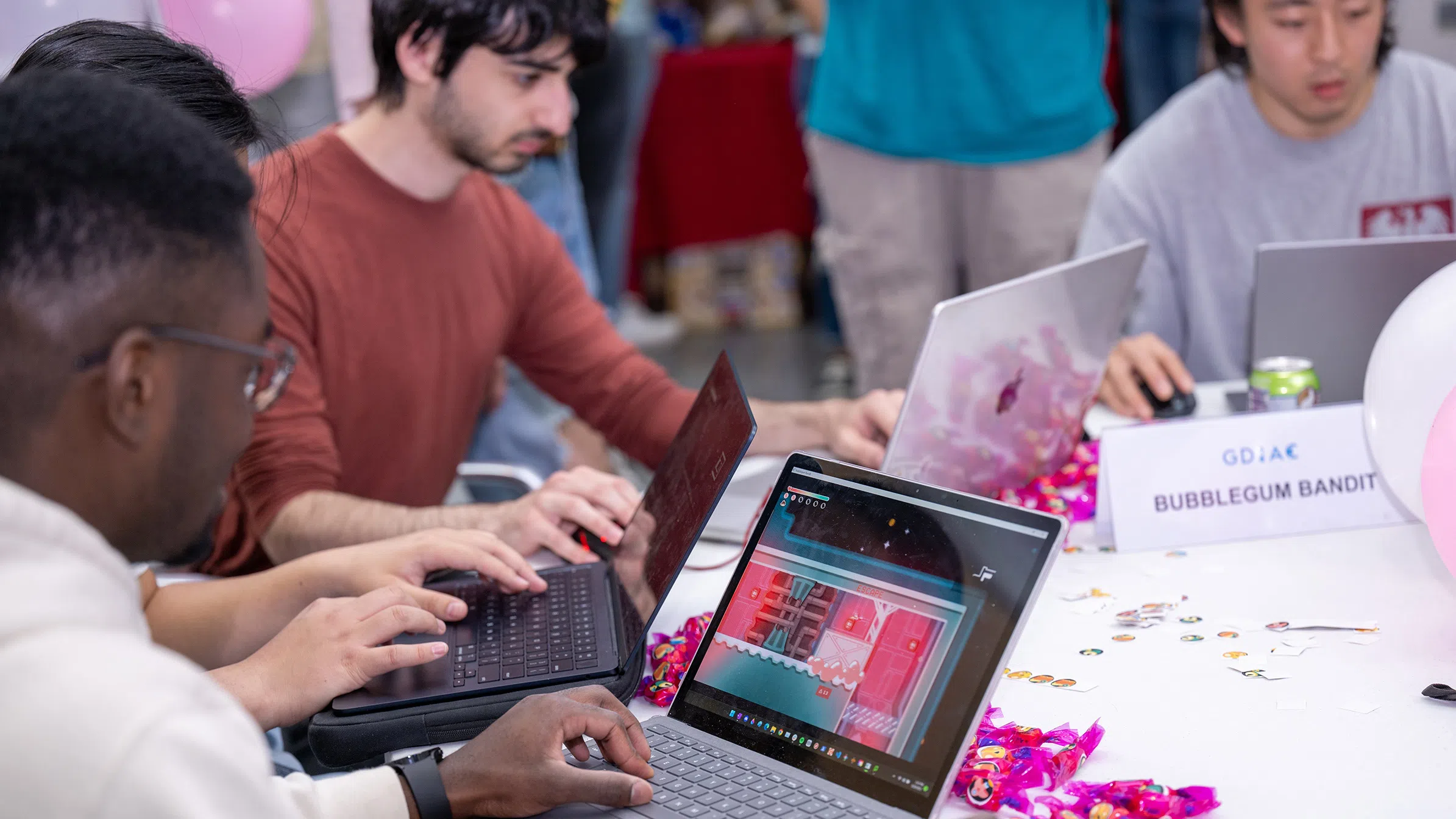 Three students work on laptops at a table. On the table is a small sign that reads “Bubblegum Bandit.”