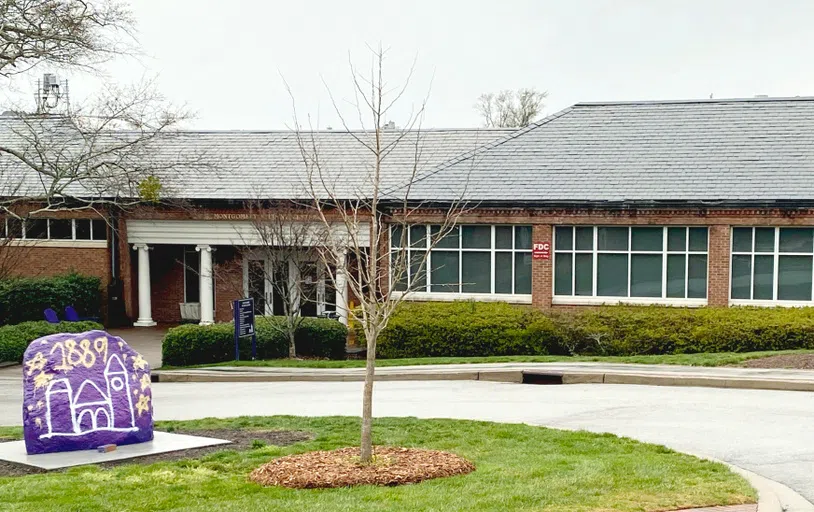 Entrance to brick building with covered portico and two sets of double doors. A large painted rock featuring purple paint and the number 1889 sits to the left in the foreground.