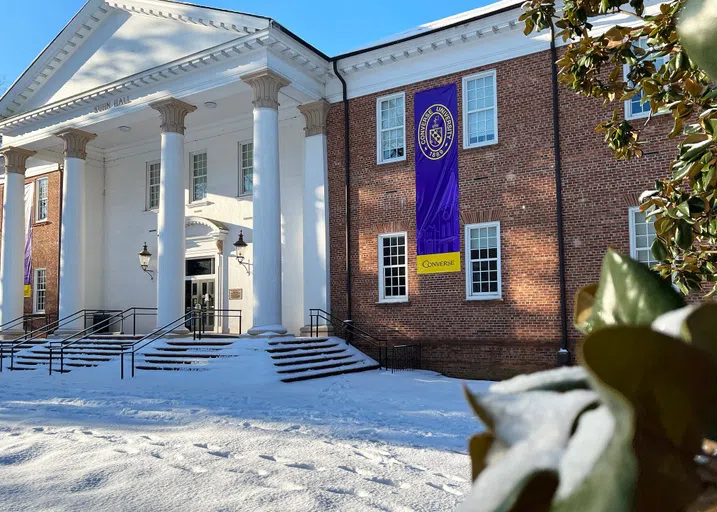 Slightly offset view of two story brick building with four-column white portico. Snow blankets the stairs and walkway leading up a black double door.