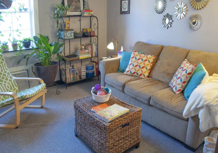 Comfortable room with sofa and rocking chair. Several plants line the wall beneath the window.