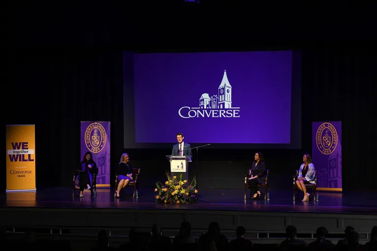 A man on stage speaks at a podium in front a purple screen with the Converse logo in white. Two women sit in chairs on each side of the podium.