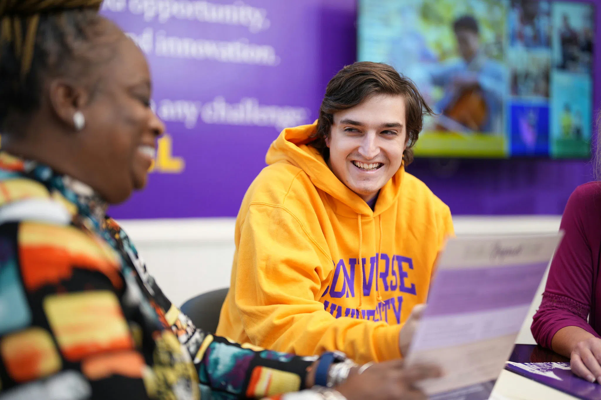 A young man in a bright yellow-orange sweatshirt smiles as a woman on the left edge of the photo in a colorful top holds out a paper in front of him.