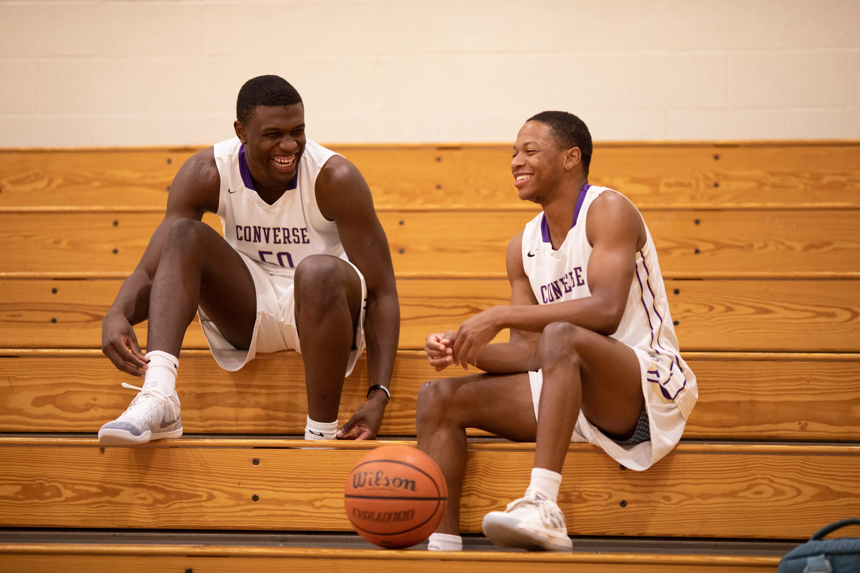 Two men sit on bleachers in white basketball uniforms with a basketball sitting between them.