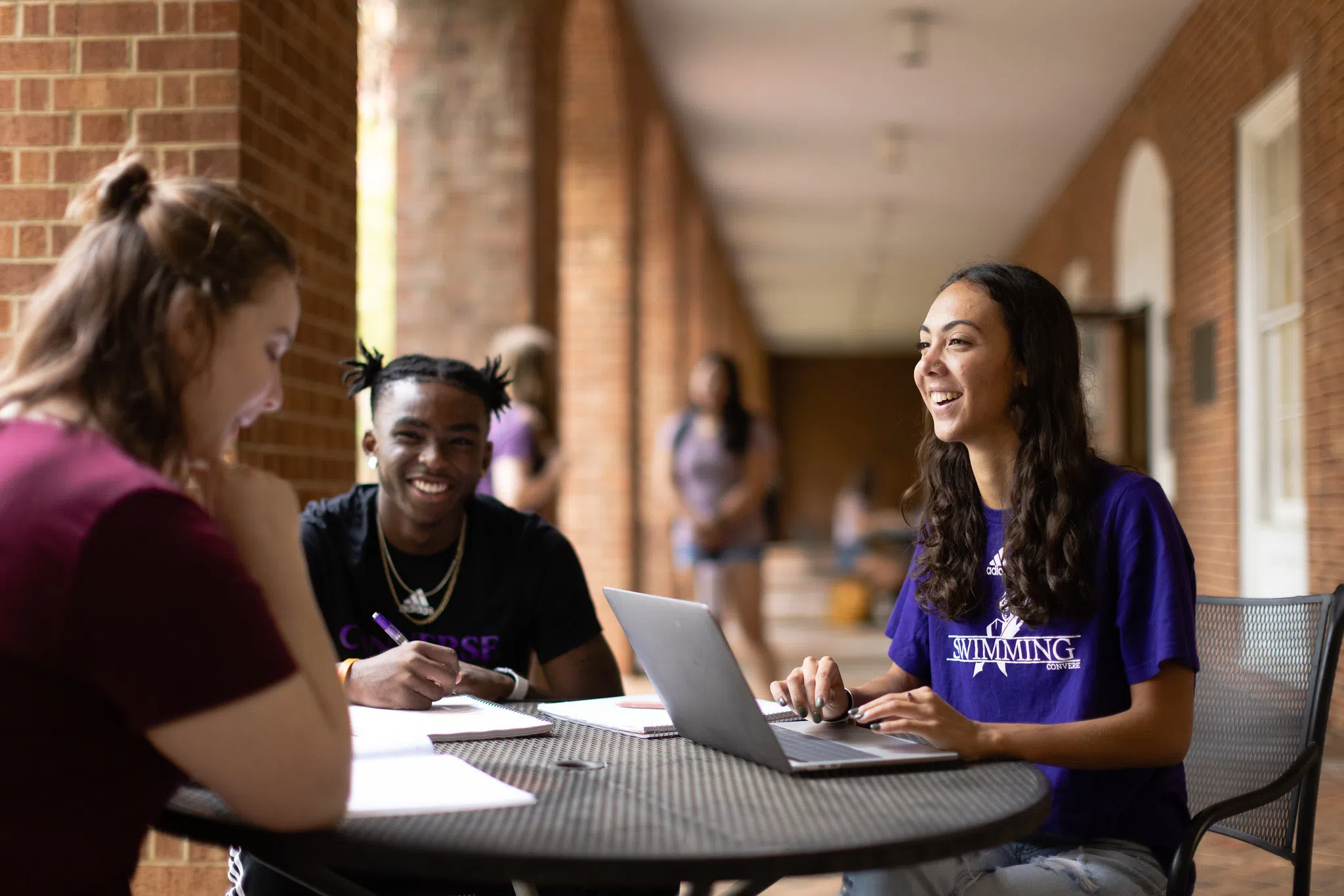 A female student with a computer sits at an outdoor table with another female student and a male student.