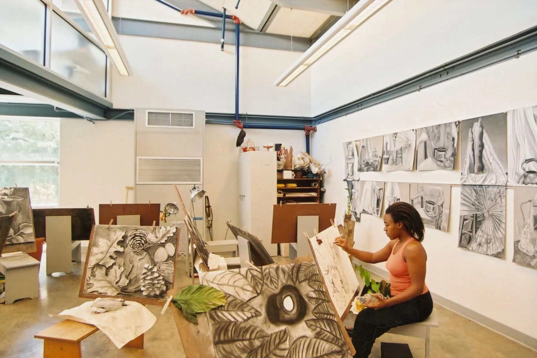 An adult female student sketches on an easel in a large white studio space surrounded by 2- and 3-dimensional art pieces.