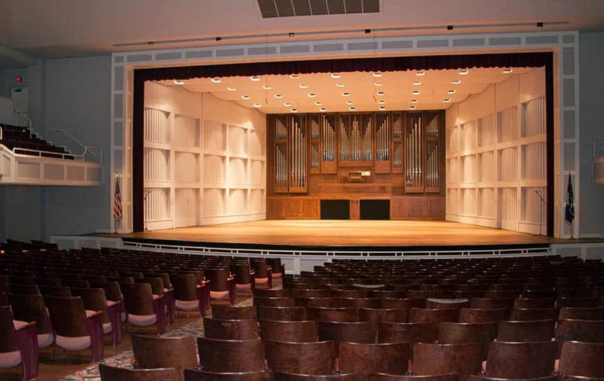 An empty stage appears from the audience view. Organ pipes can be seen set within wood panels at the back of the stage.