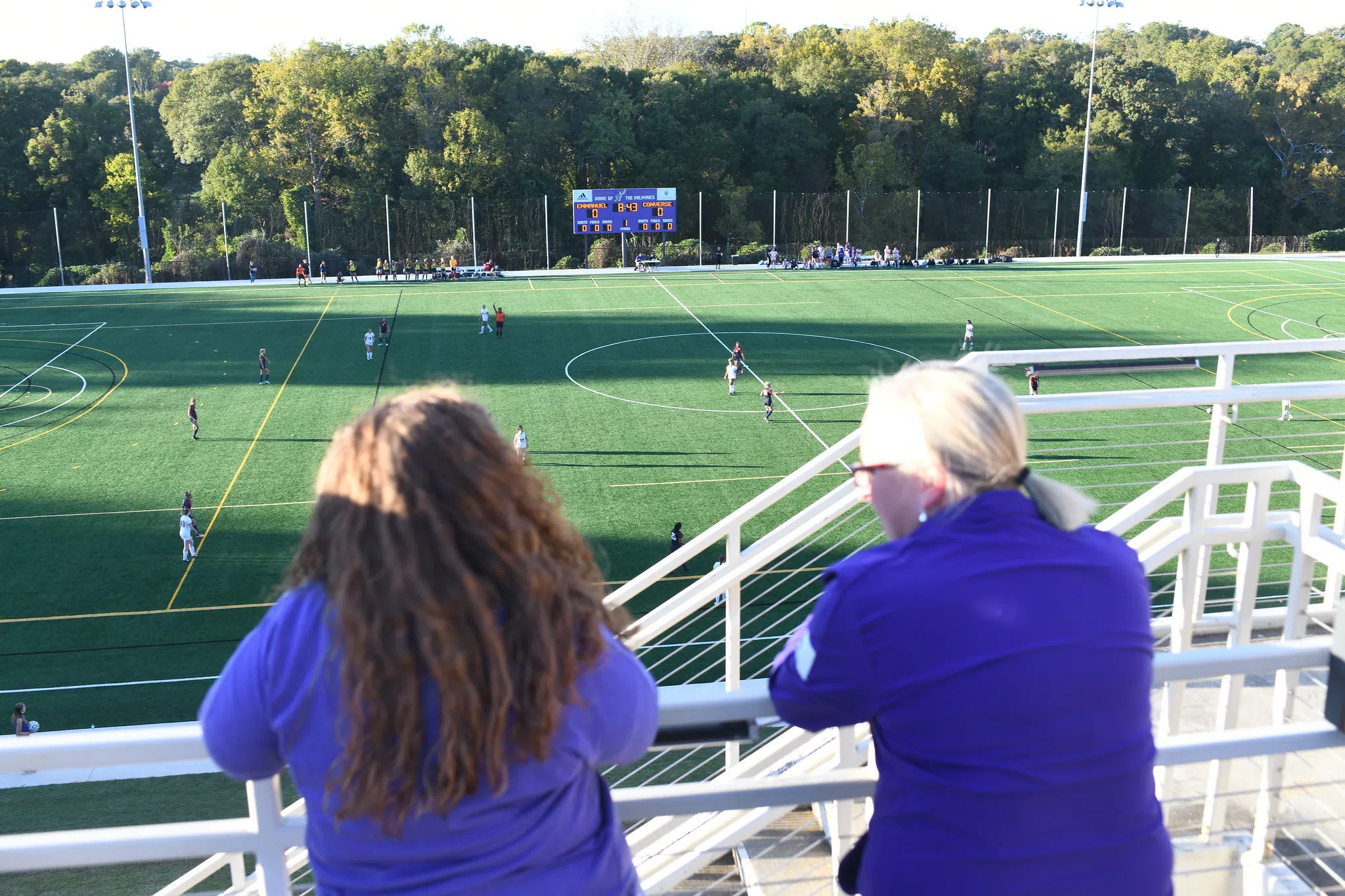 Two women with their backs to the camera survey a green athletic field below them. Distant students are scattered on the field mid-play.  