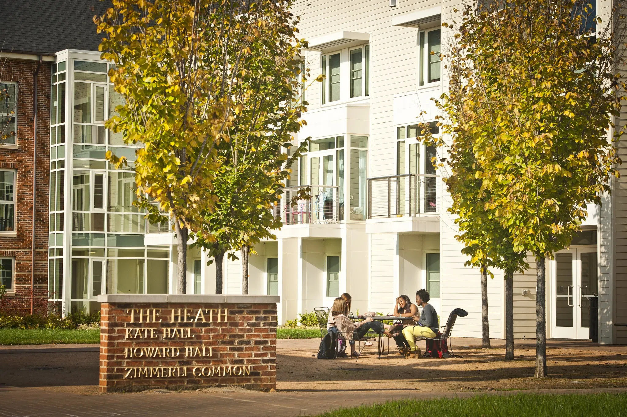 Four students sit at an outdoor table in front of a three story building. A sign in the foregrounds says "The Heath. Kate Hall. Howard Hall. Zimmerli Common.