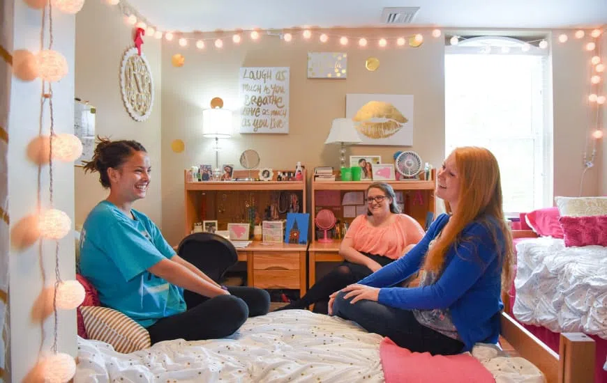 Three women sit on beds chatting in a dorm room.
