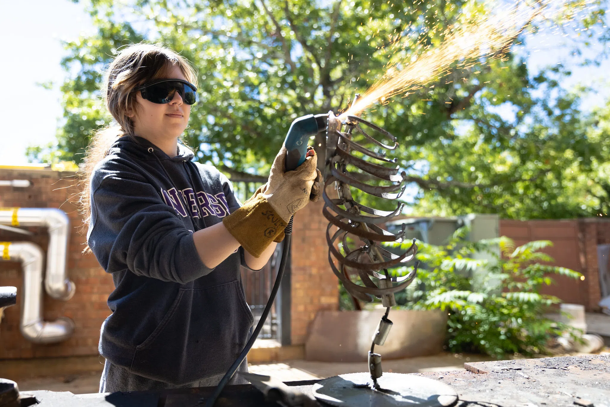 A student wearing safety goggles is outside welding a metal form as sparks shoot from the welding tool.