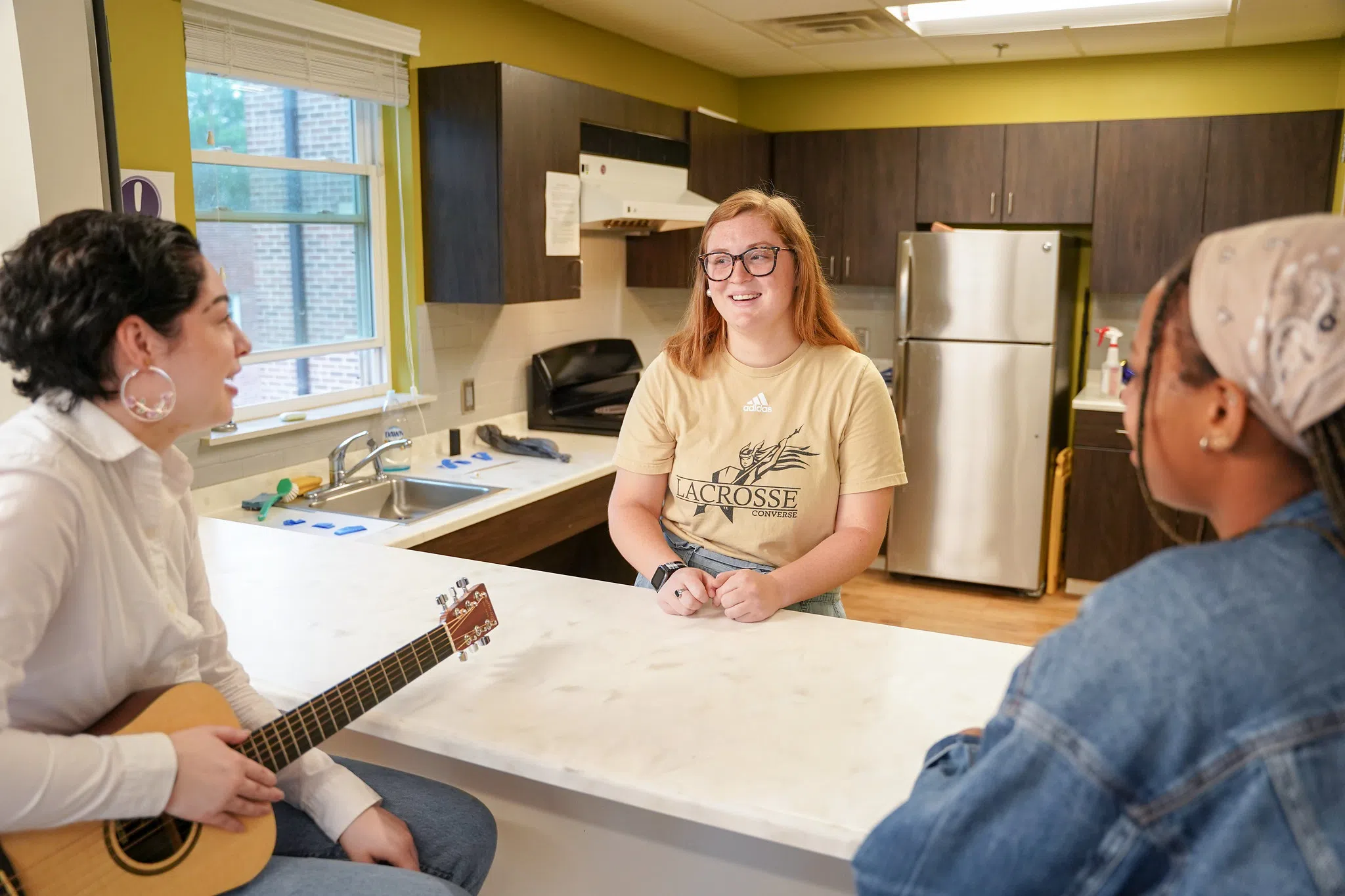 A woman in a yellow t-shirt stands on one side of a kitchen counter speaking with two other women, one holding a guitar, on the other side of the counter. A stovetop and refrigerator can be seen in the background.