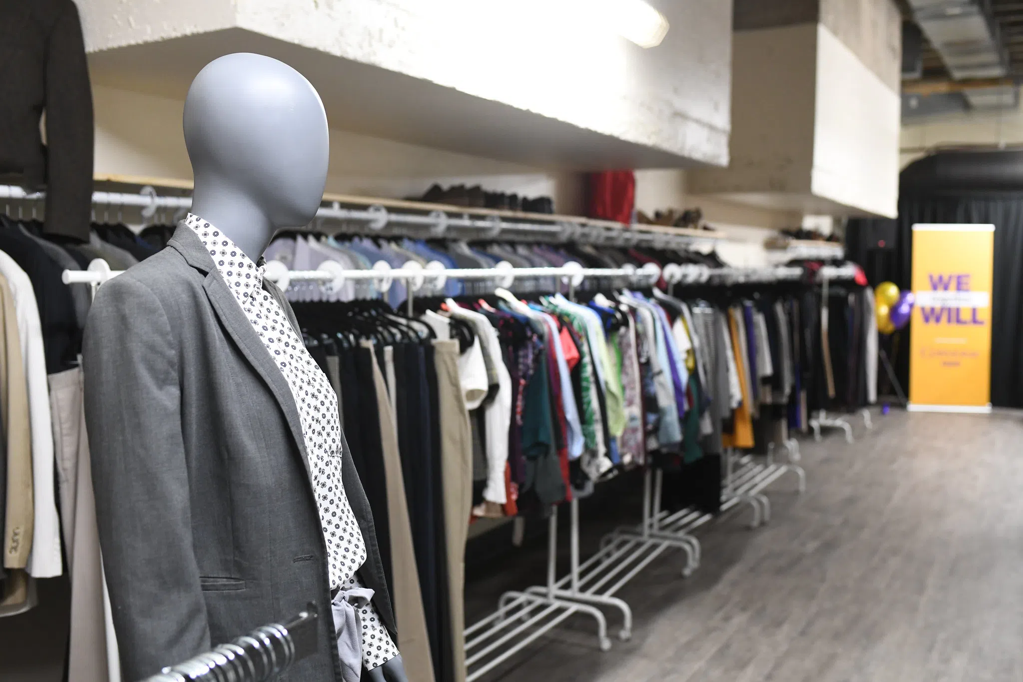 Clothing racks with hanging items against the wall. Mannequin dressed in business casual outfit displayed out front. 
