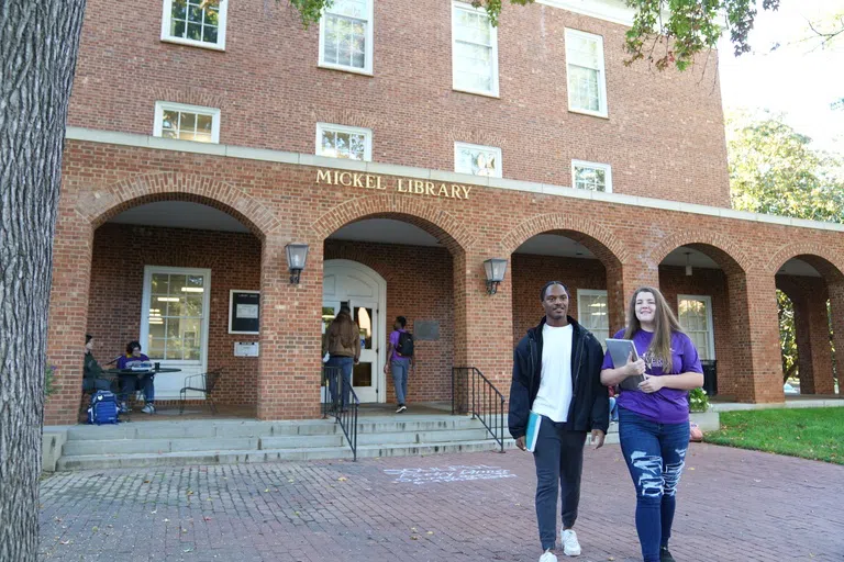 Two students in the foreground walk away from the entrance of a three story brick building featuring five brick arches leading to a covered porch. The words "Mickel Library" appear in gold letters above the arches. In the background, two students enter the building and two students sit at a black iron table.