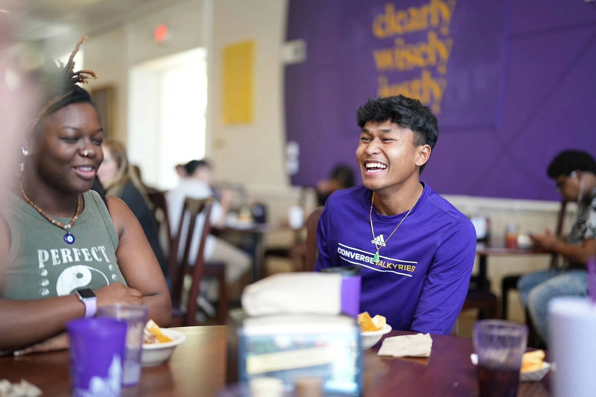 A smiling student with dark hair and a bright purple tshirt laughs with others around a table in the dining hall.