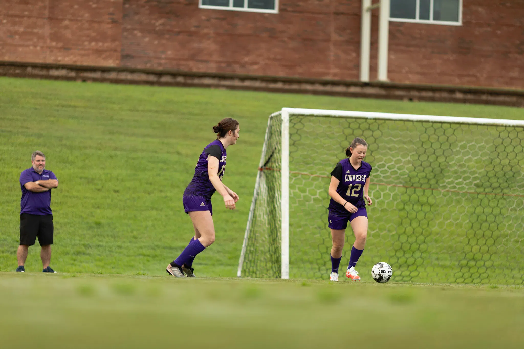 Two female players in soccer uniforms kick a ball near a large soccer goal. A male coach looks on from the sidelines. A portion of a brick building appears in the background.