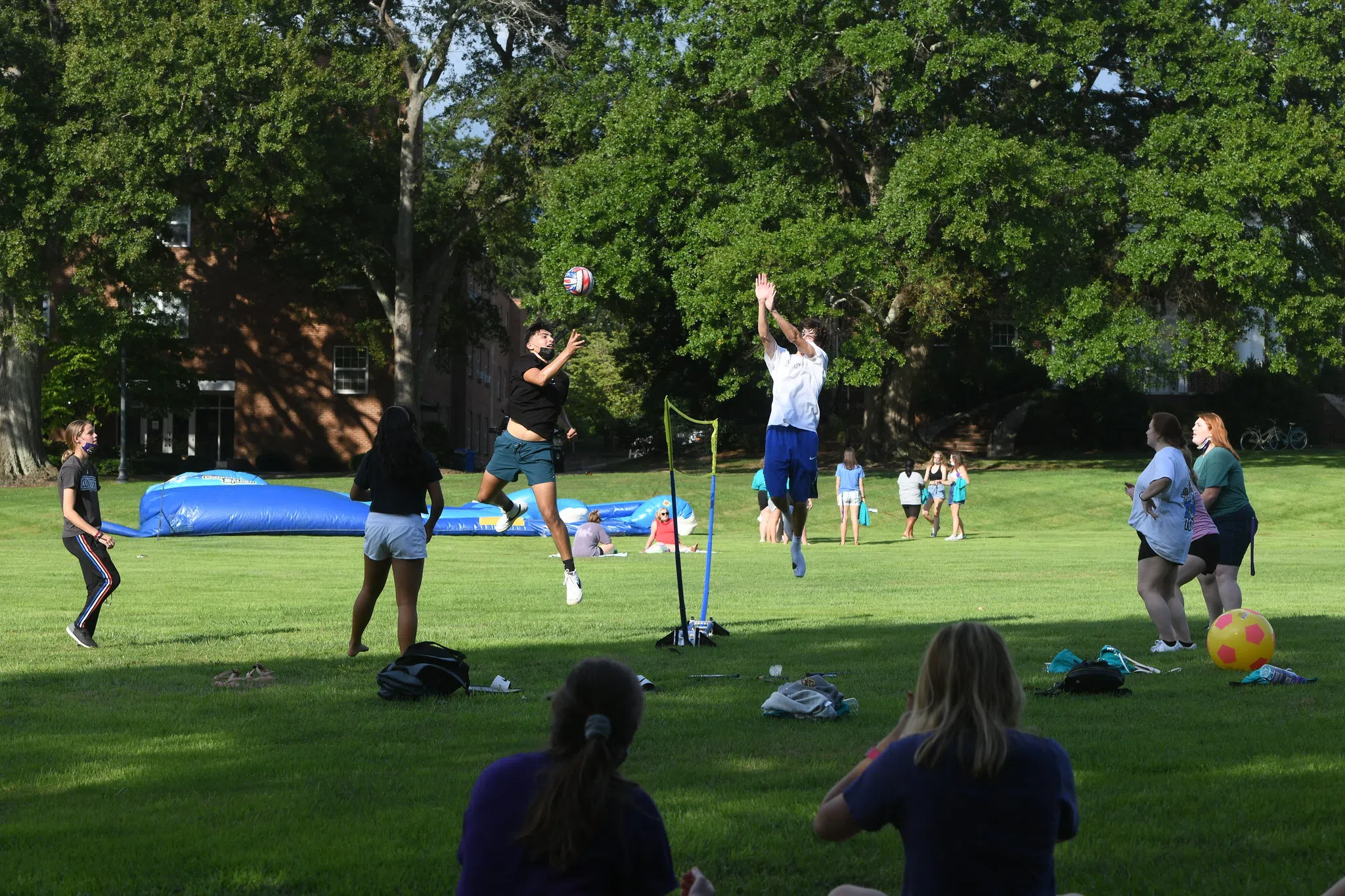 Male and female students play a game of volleyball in a grassy, open field on a sunny day. A large inflatable game is being set up in the background.