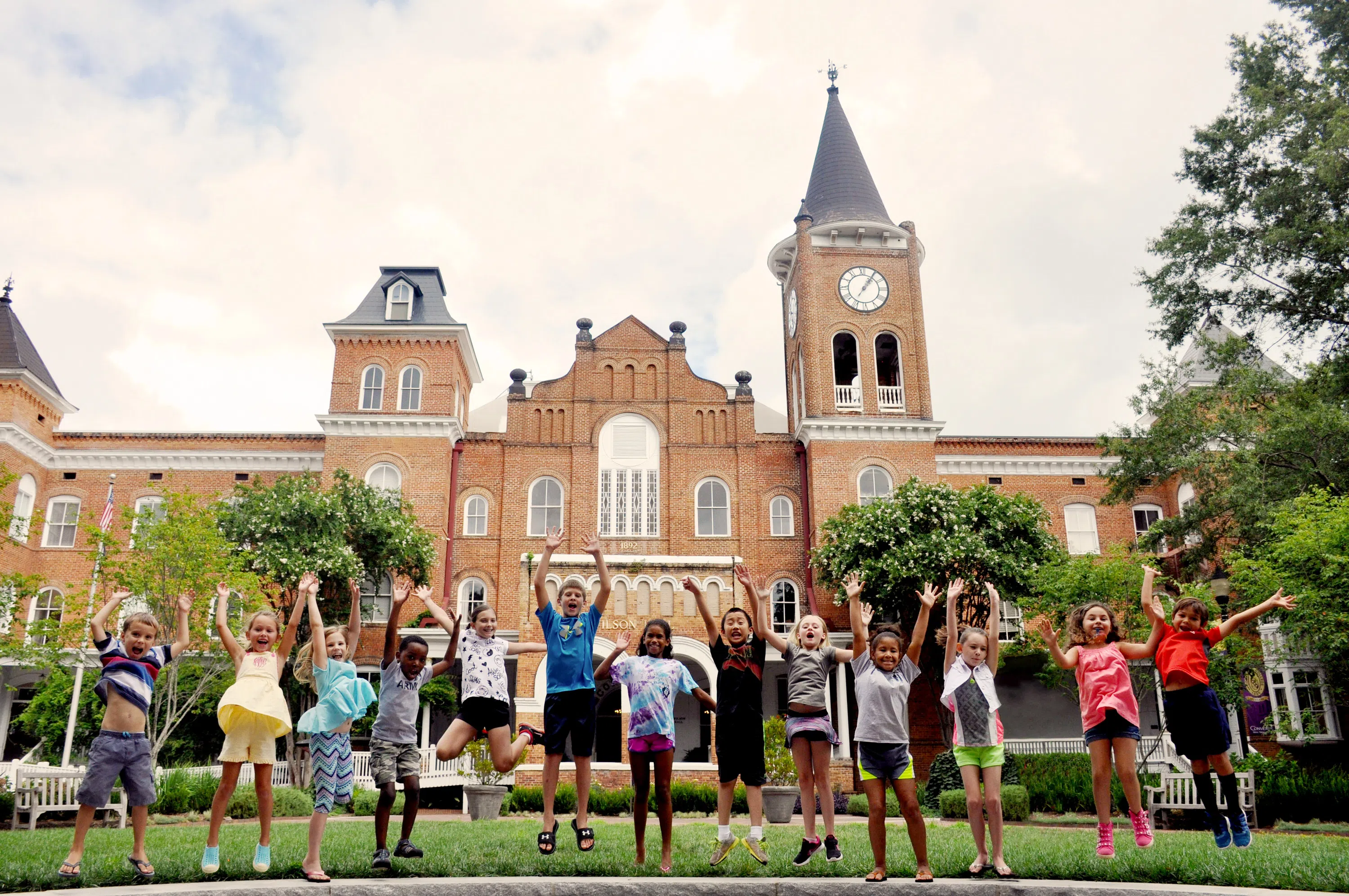 Elementary age students jump in the air in a forward facing line while enjoying a sunny day an outdoor space. A large brick building appears in the background.