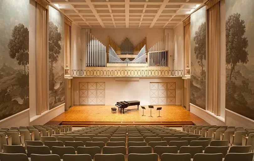 A piano, a black leather bench and three music stands appear on a stage below wall-mounted organ pipes. Walls featuring murals showing outdoor scenes surround the stage.