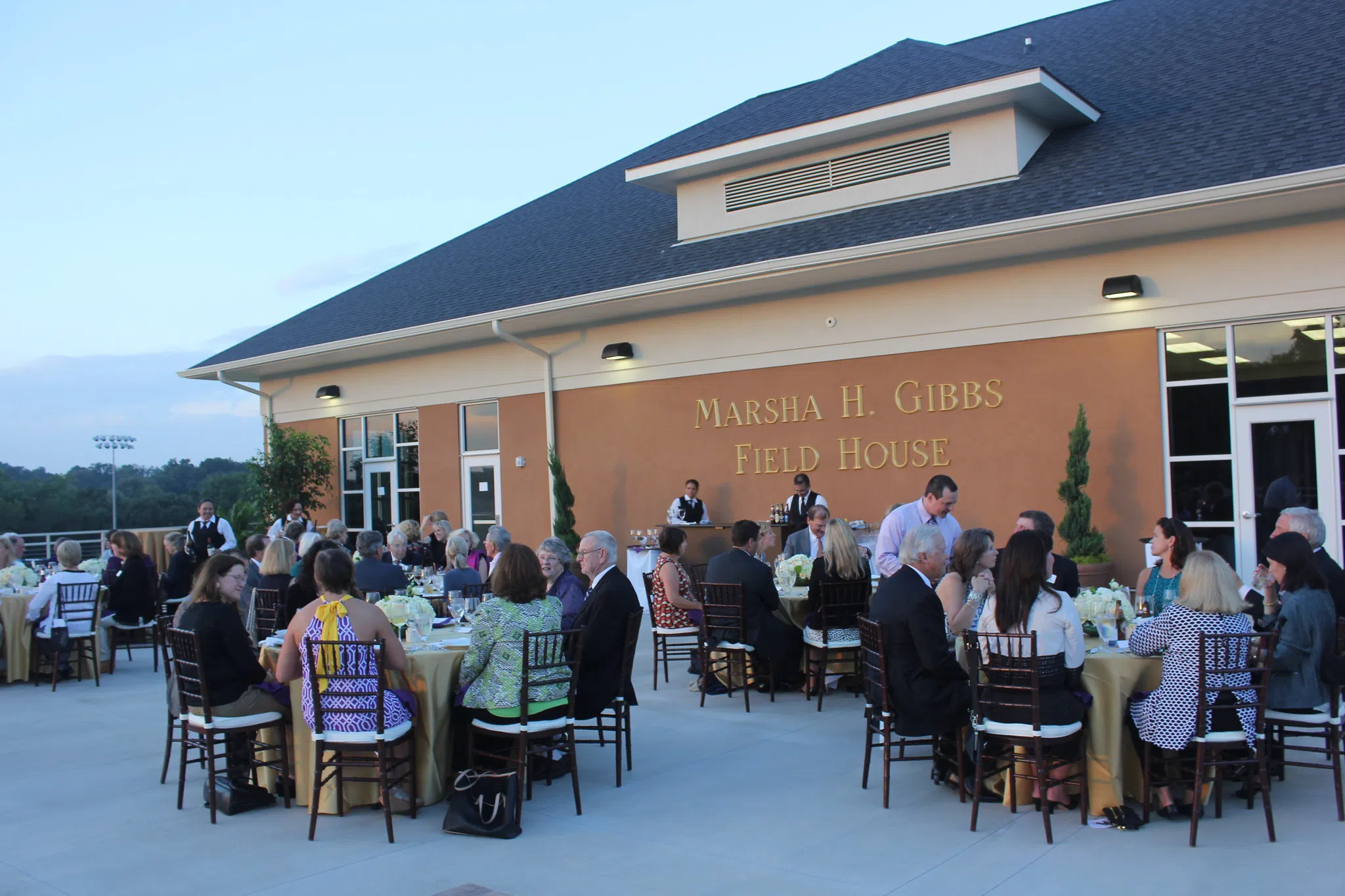 Guests dine at outdoor tables with gold chairs and cloths. A brick wall in the background reads: Marsha H. Gibbs Field House.