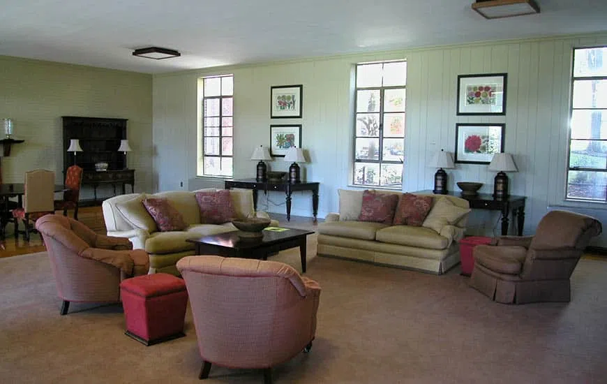 A pale-wood paneled room with couches and arm chairs and three windows separated by framed wall prints.