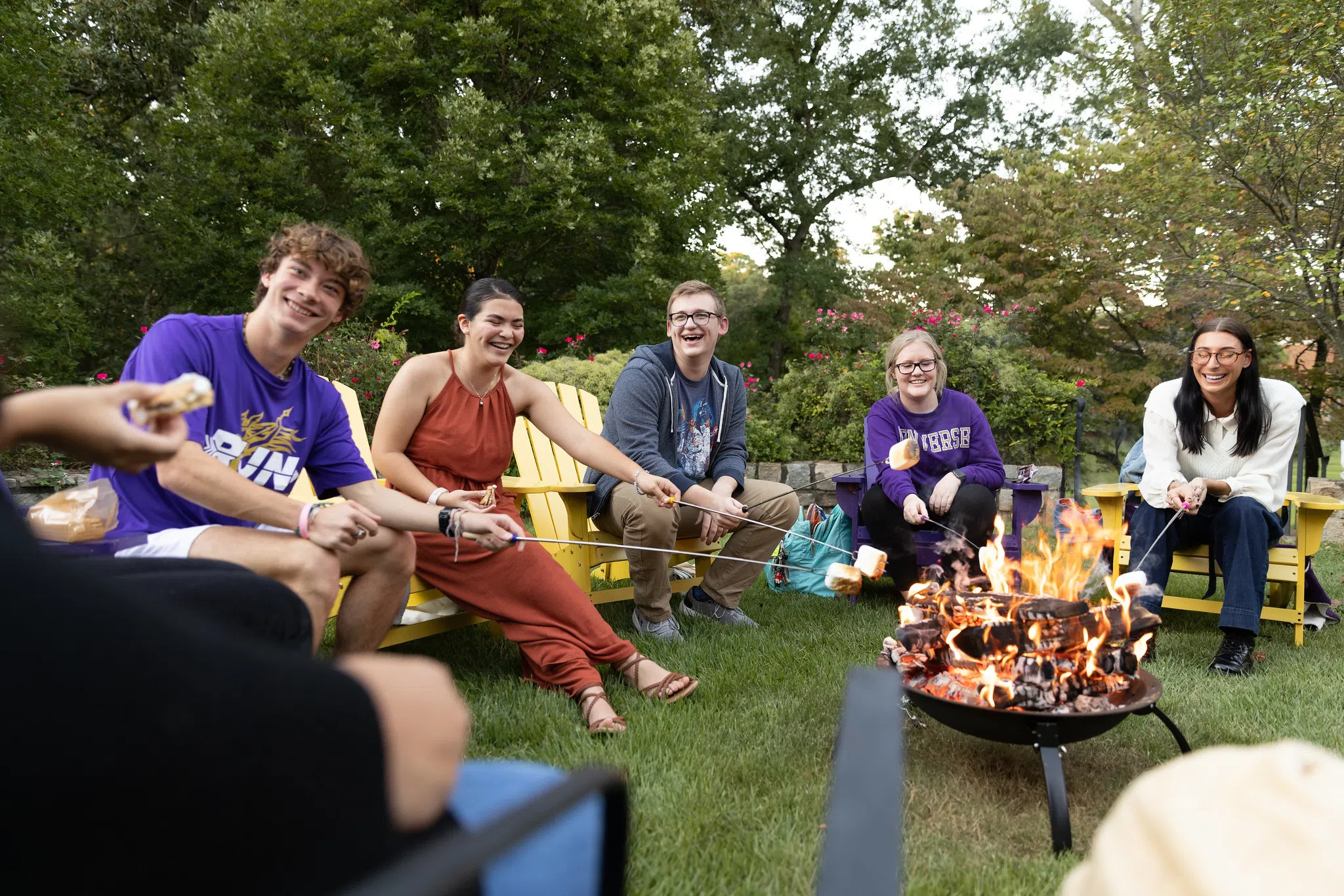 A group of students sit in Yellow Adirondack Chairs with sticks bearing marshmallows. A large fire blazes in a metal fire pit to the right.