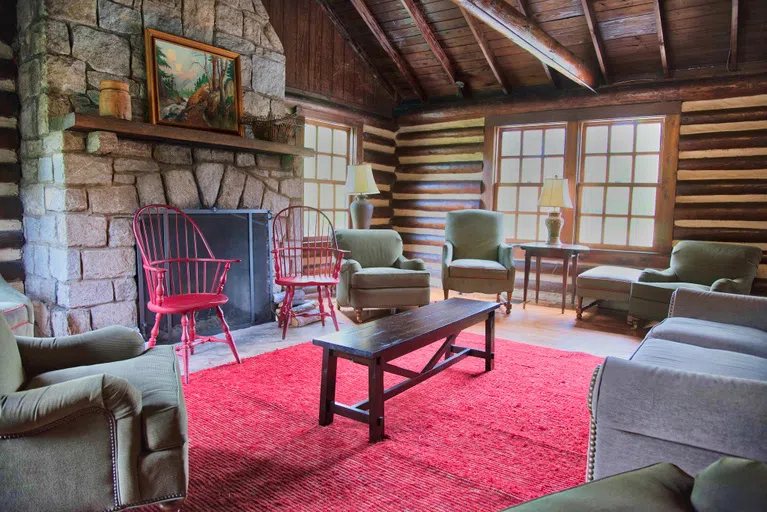 A variety of small tables, sofas and chairs appear on a red rug, facing a large stone fireplace. Wooden wall and large windows appear in the background.