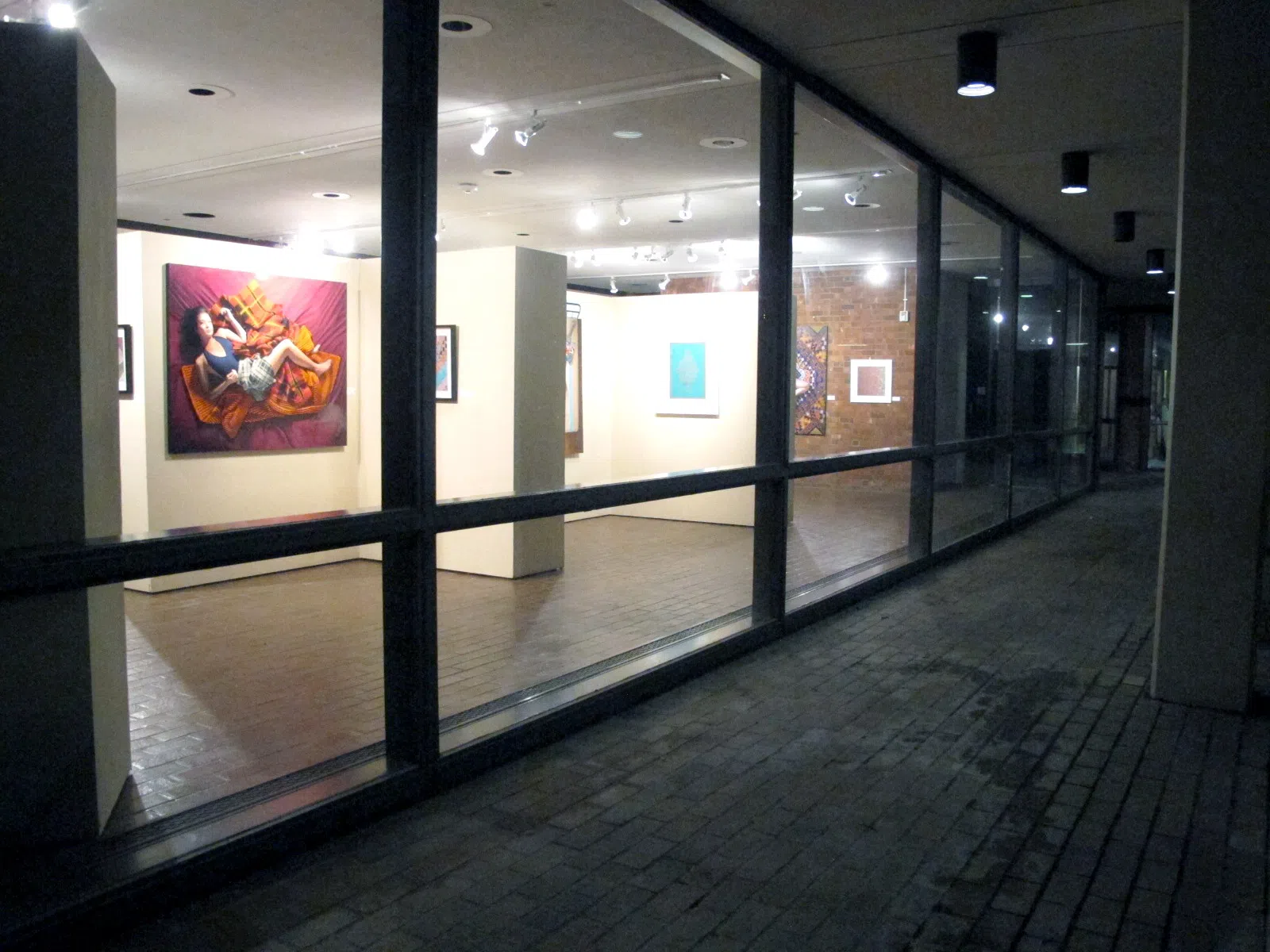 A well-lit art gallery is visible behind windows lining an outdoor brick path at night.