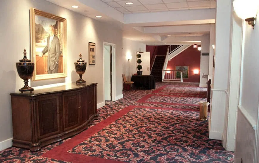 Art work, tables and an urn line the left side of a red-carpeted room. Stairs appear in the background.