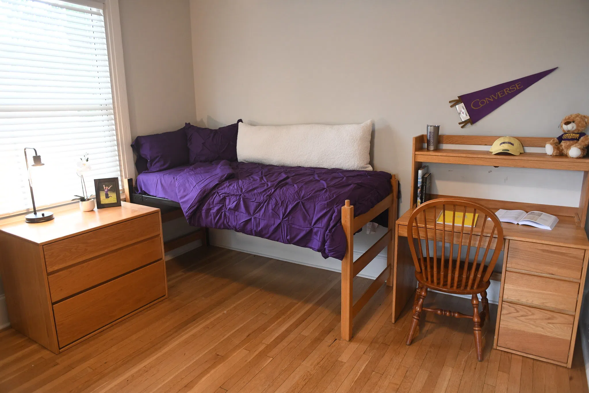 A lofted bed with purple blanket and pillow appears in a corner between a desk on the left and a bedside chest with three drawers below a window on the right.