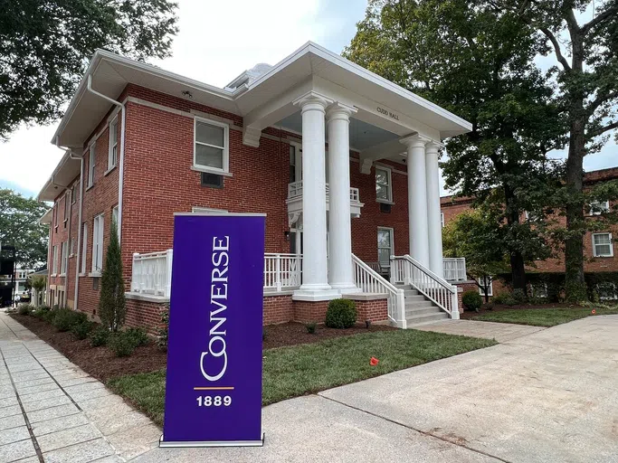 Main entrance to three-story Cudd Hall. Four white pillars support a patio on the brick building. A purple sign in the foregrounds shows "Converse 1889".