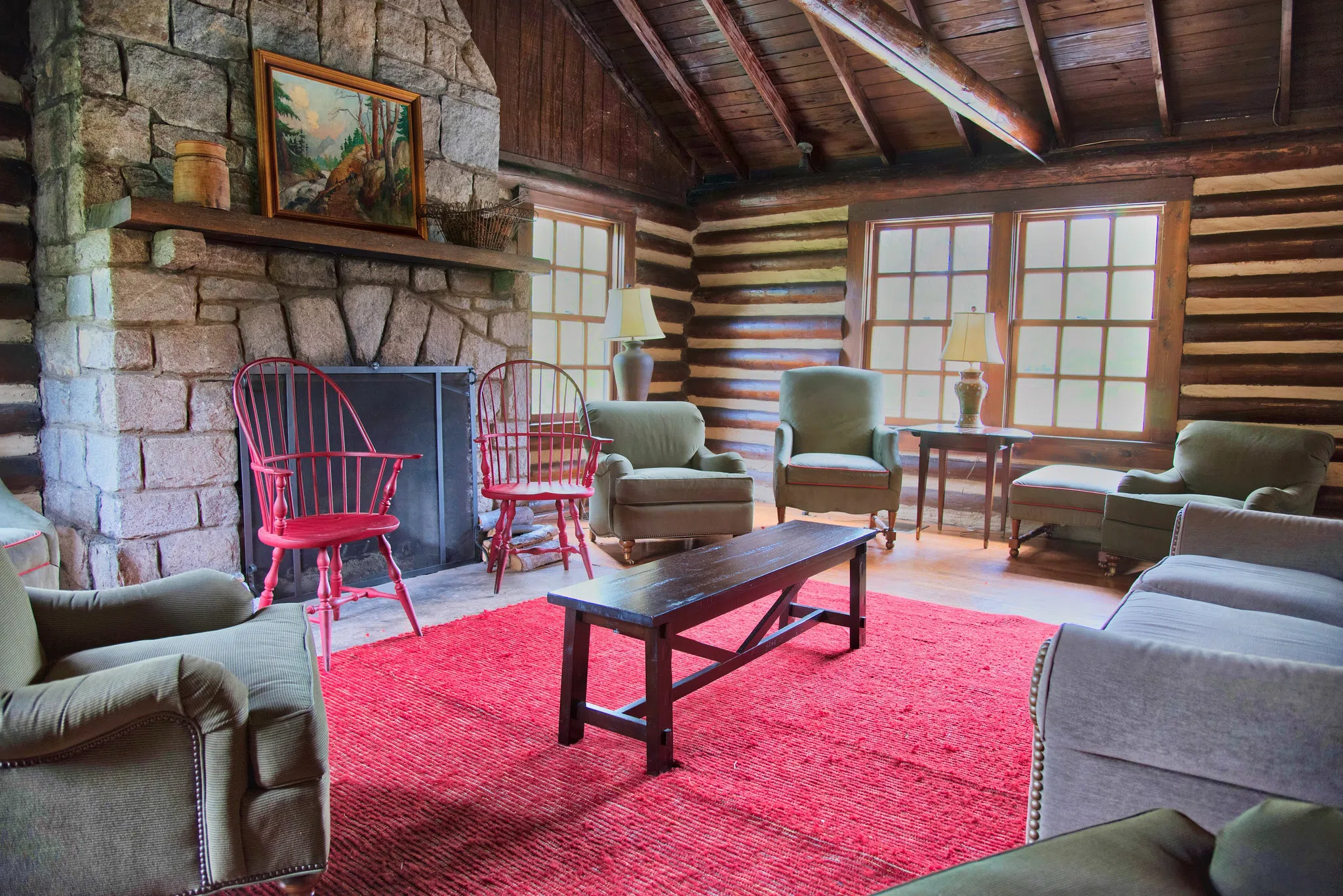 A variety of small tables, sofas and chairs appear on a red rug, facing a large stone fireplace. Wooden wall and large windows appear in the background.