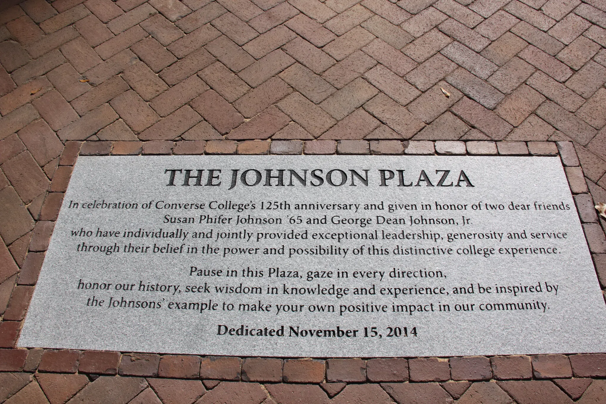 A gray concrete plaque set within a brick walkway is engraved with the words "The Johnson Plaza - In celebration of Converse College's 125th anniversary and given in honor of two dear friends Susan Phifer Johnson ’65 and George Dean Johnson, Jr. who have individually and jointly provided exceptional leadership, generosity and service through their belief in the power and possibility of this distinctive college experience. Pause in this Plaza, gaze in every direction, honor our history, seek wisdom in knowledge and experience, and be inspired by Johnson’s example to make your own positive impact in our community. Dedicated November 15, 2014