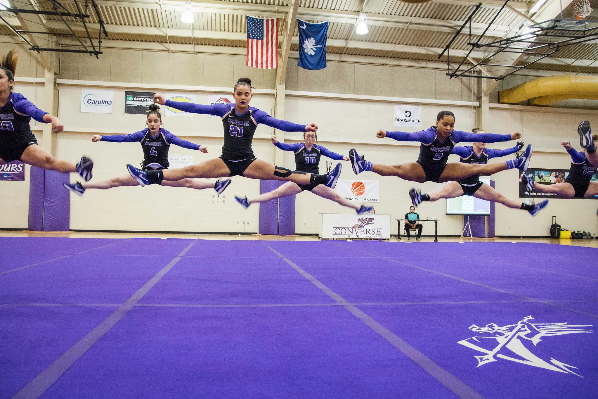 Athletes from the Acrobatic and Tumbling team perform straddle jumps on a purple gym mat.