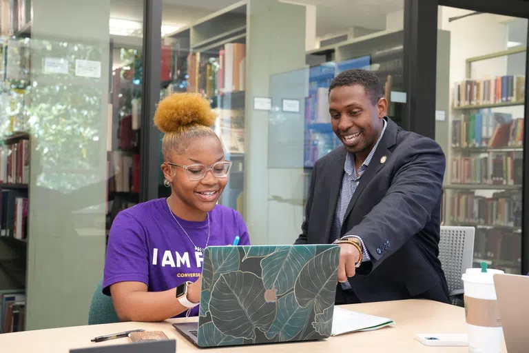 A man in a gray suit sits at a table and points to a computer screen as a student in a purple shirt that reads "I Am First" looks at the screen with a smile. A wall of glass windows separate them from shelves of books in the background.