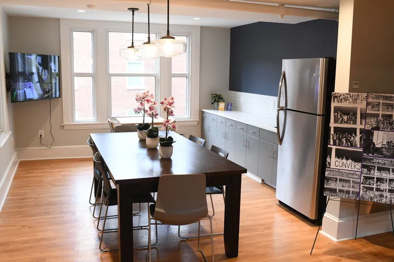 A kitchen area features a table with six chairs in front of a large window, alongside a counter with cabinets and a sink. A stainless steel refrigerator appears in the right foreground.